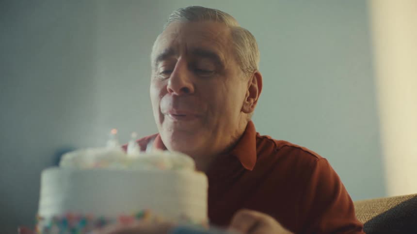 Man holds a birthday cake with lit candles as he blows out the candles and kisses his wife sitting next to him
