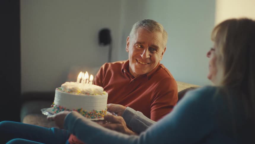 An older man is smiling as he blows out the candles on a birthday cake held by an older woman