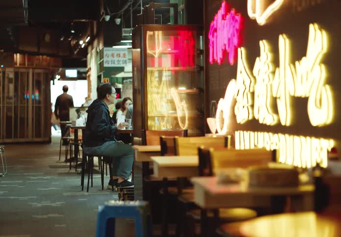 A man seated indoor at the restaurant table with people walking around