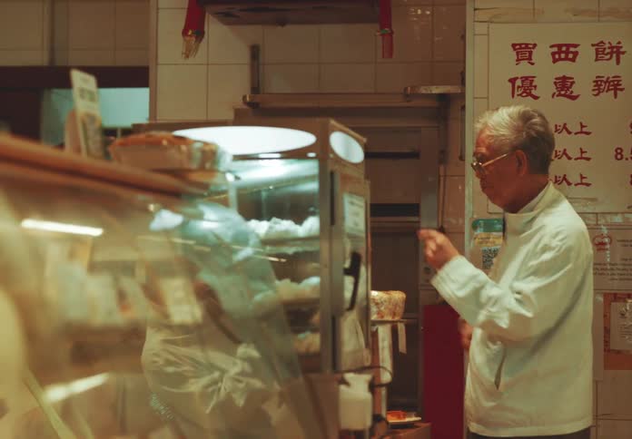 Elderly man serves food in a traditional chinese bakery next to a glass counter and a menu board in mandarin