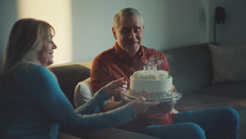 Woman holds a birthday cake with lit candles on it up to a man who blows the candles out