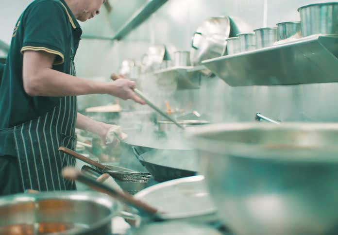 A chef is cooking and transferring food from a pan into a strainer with a younger chef standing beside him assisting in the kitchen