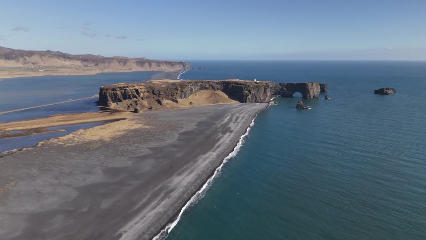 An aerial view captures a black sand beach meeting the turquoise ocean alongside a rocky coastline and cliffs under a clear blue sky