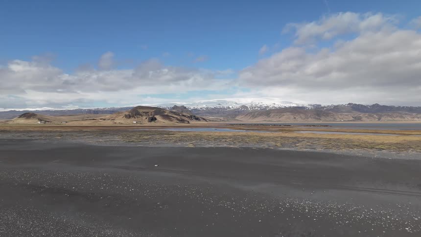 Aerial shot of a beach with a long stretch of black sand and snowy mountains in the background