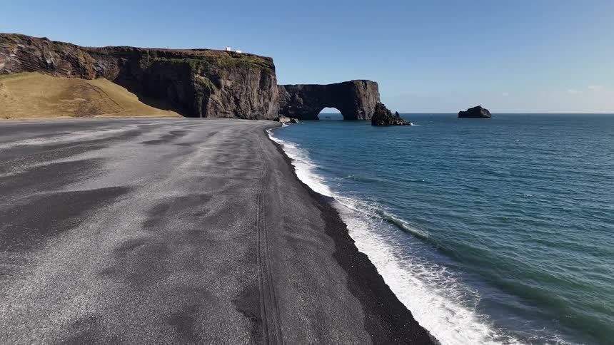 Waves crash onto a gray sandy beach bordering a promontory with a natural arch