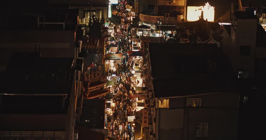 Aerial view of a crowd of people moving through a bustling street market at night with colorful lights illuminating the stalls and walkways