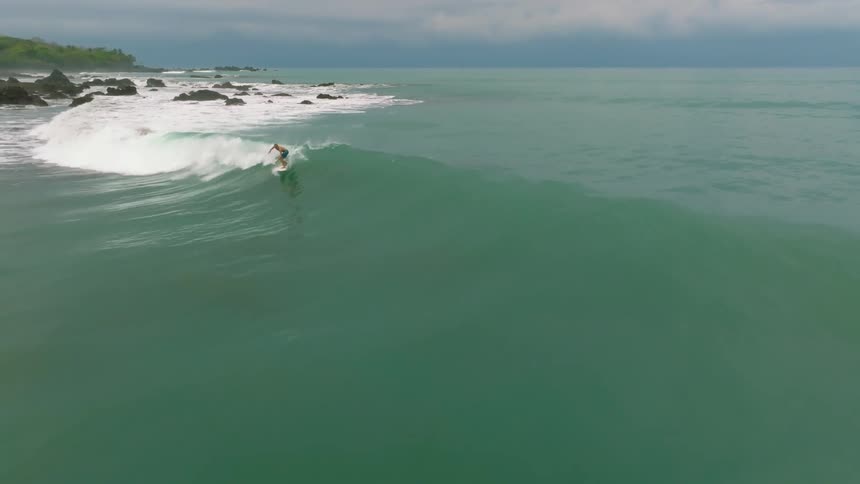 A lone surfer rides a curling wave near a rocky shoreline under an overcast sky
