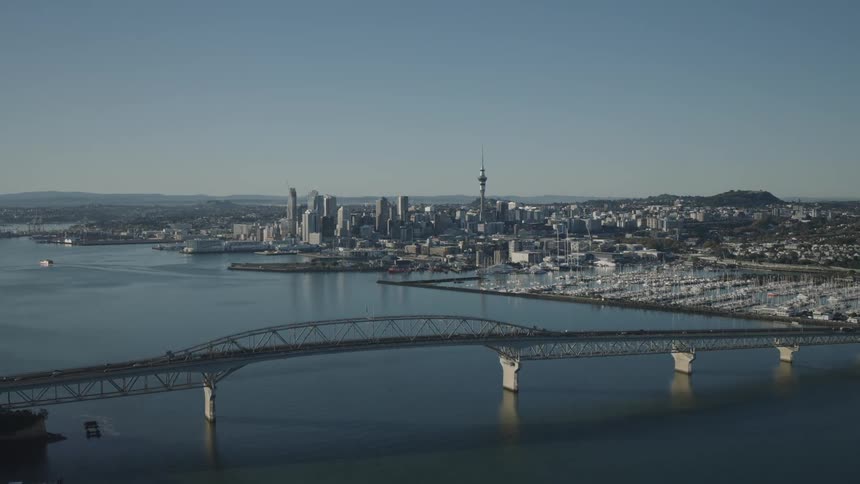 Auckland harbour bridge spans the calm waters leading towards the city skyline dominated by the sky tower with numerous boats docked in a marina to the right