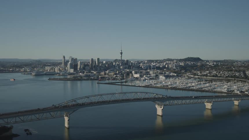 A wide aerial shot captures aucklands city skyline featuring the iconic sky tower and harbour bridge over calm blue water