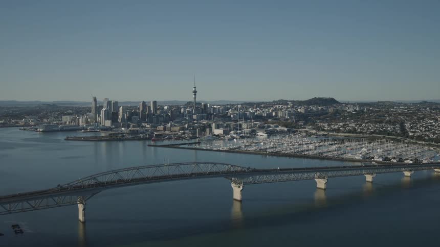 Auckland harbour bridge spans the calm waters leading towards the city skyline dominated by the sky tower with numerous boats docked in a marina to the right