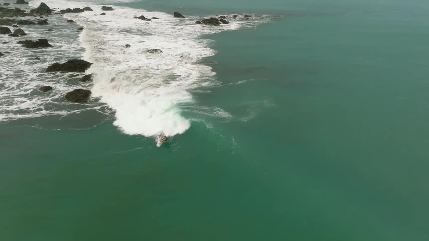 A surfer rides a wave along a rocky coastal shoreline in an aerial view