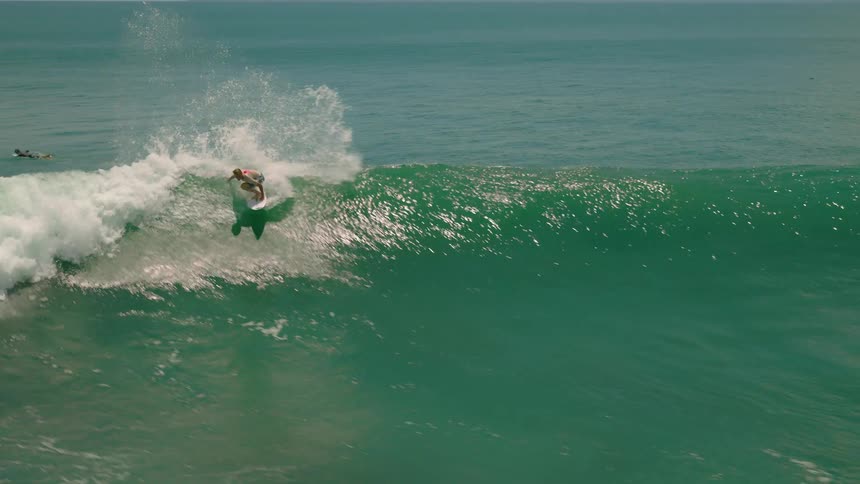 A surfer skillfully rides along a sunkissed green ocean wave on a surfboard performing tricks before losing balance and falling into the churning whitewater