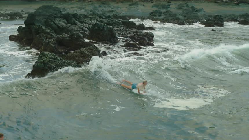 A man on a beach standing on a rock in the sea jumping onto a surfboard
