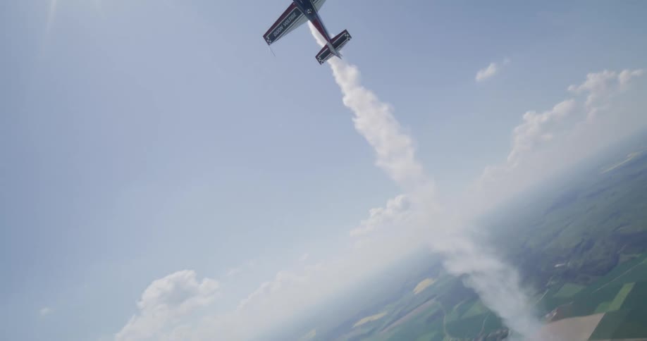 A small airplane performing aerial maneuvers trails white smoke across a landscape of patchwork fields villages and a distant highway under a mostly clear blue sky