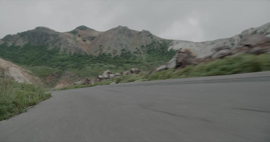A car drives along a mountain road surrounded by rugged greenandgray hills under overcast skies