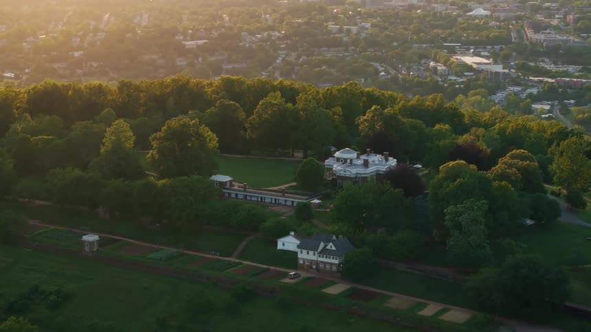 Monticello stands amidst lush trees and cultivated gardens with a cityscape visible in the hazy distance bathed in the warm glow of either sunrise or sunset