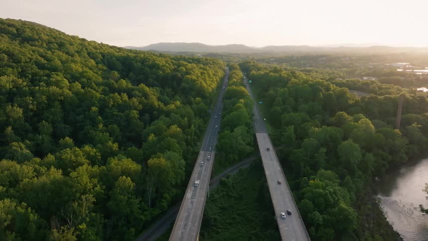 An aerial view captures cars and trucks traveling along two parallel highway bridges that cut through a dense green forest alongside a river with distant mountains visible