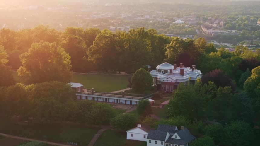Groves of trees surround large buildings located on a hill overlooking a residential area while the sun tints the fog red
