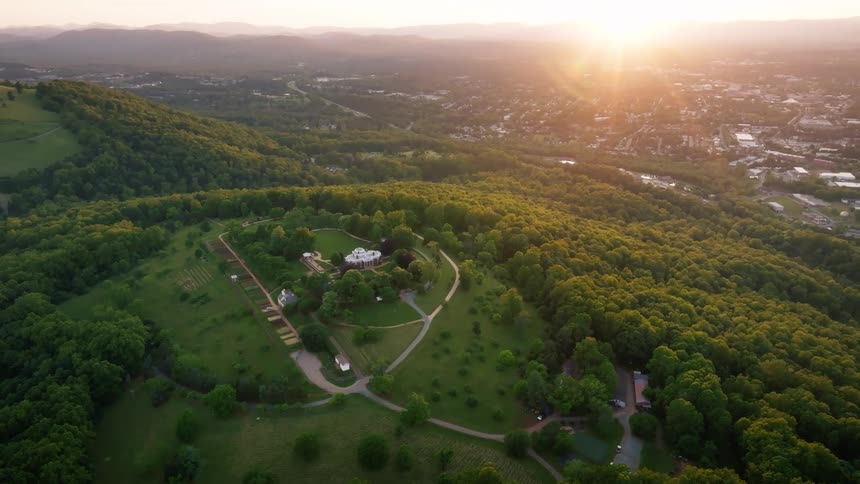 An aerial shot captures a sprawling estate with a central mansion on a forested hilltop as the sun rises illuminating a distant town and landscape