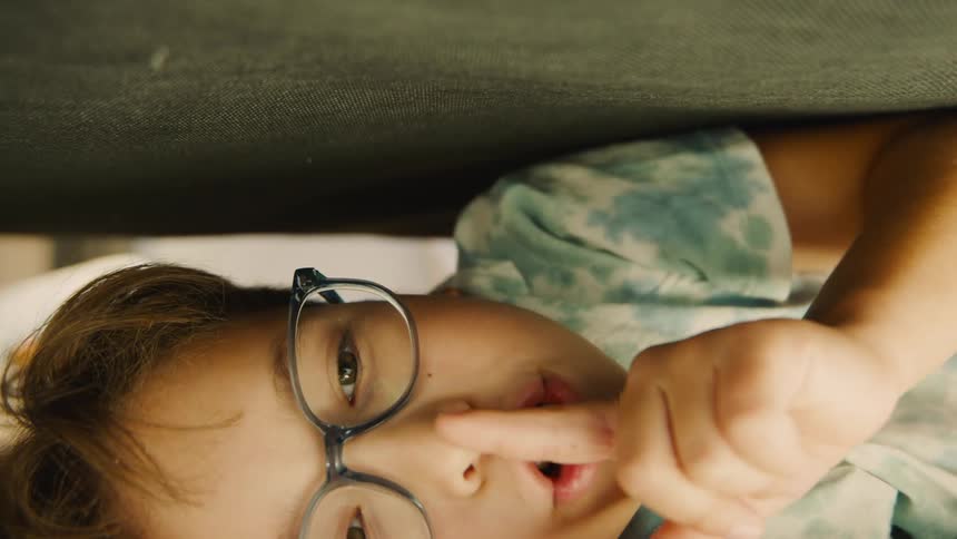 A boy wearing glasses lies on his back under the bed holding a finger to his mouth as he hides during a game of hide and seek