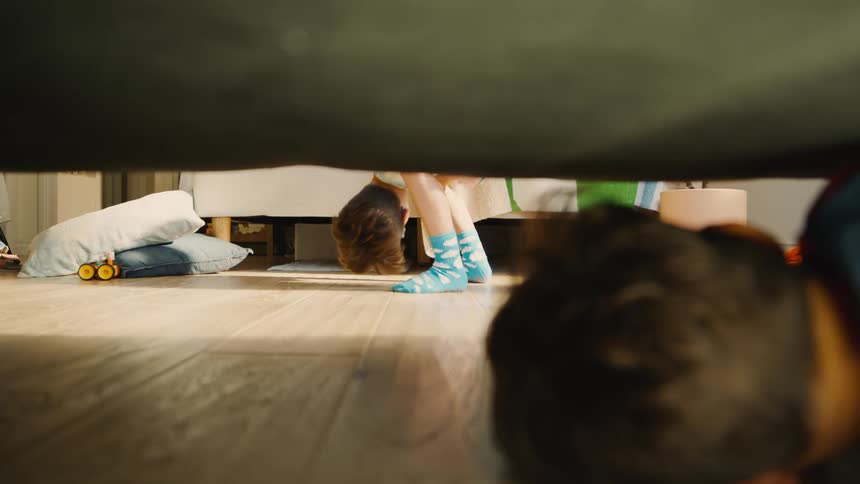 Two children playing in a room one is hiding under the bed and the other is looking for them