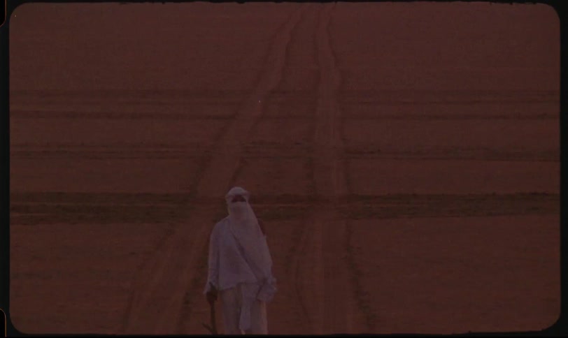 A figure draped in white clothing obscuring their features walks along a sandy track in a desolate dusky environment while holding a stick