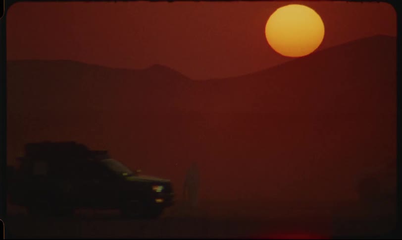 A person stands beside vehicles in a dusty desert landscape with the sun setting over distant mountains