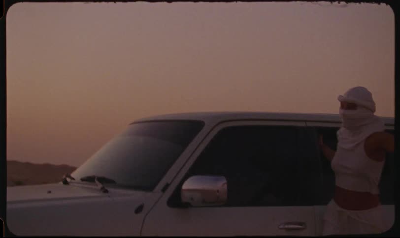 Woman wearing a head and face covering rides on the outside of a car as the sun sets over the desert