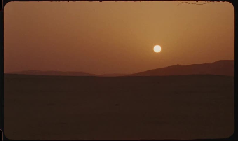 The sun dips below a stark desert horizon casting a warm hazy glow over the undulating sand dunes