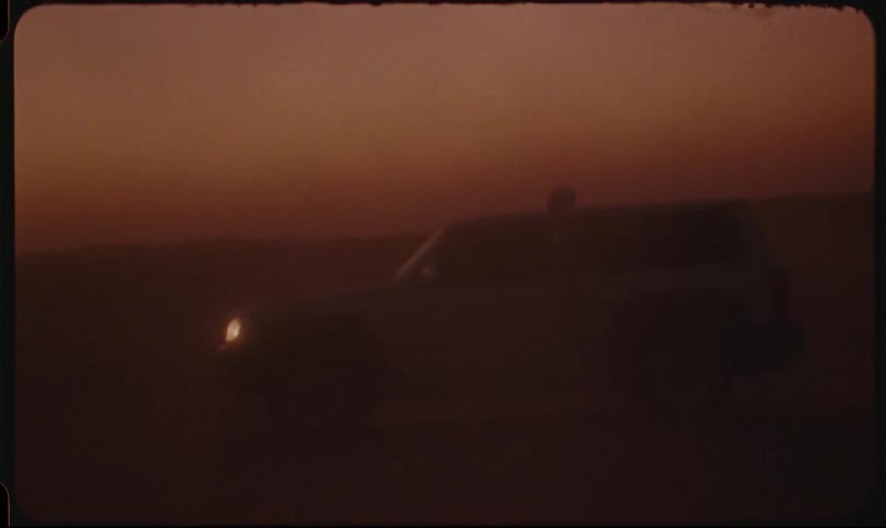 Headlights illuminate a vehicle moving across a dimly lit arid landscape kicking up a dust cloud under a dusky sky