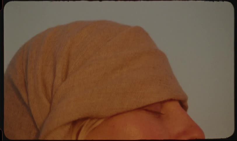 A closeup shot shows a woman with fair skin and light blonde hair having a light brown fabric wrapped around their head by someone with darker skin