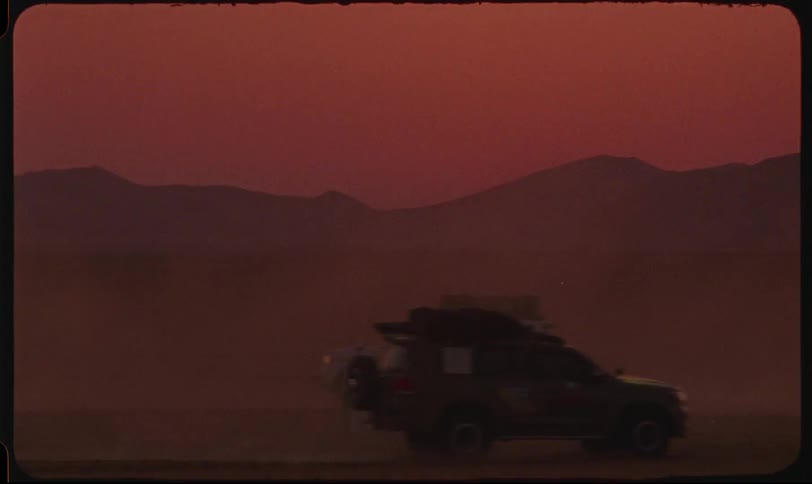 A person walks through a dusty desert while three cars drive past at sunset