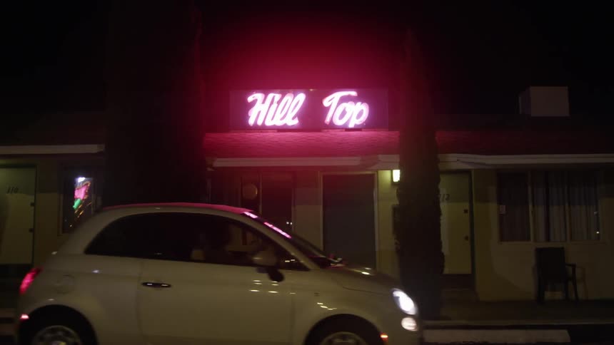 A car pulls up in front of a building with a neon sign
