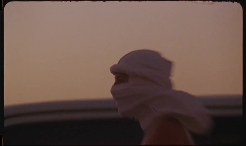 Woman wearing a headscarf leans against the window outside a vehicle moving through the desert at sunset