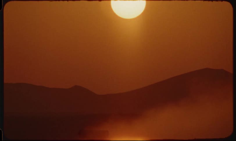 A truck driving through the desert sand at sunset with dust rising into the air