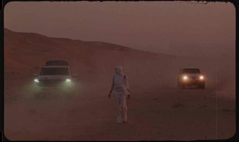 Woman in a white dress walking through the desert and vehicles passing at high speed around her