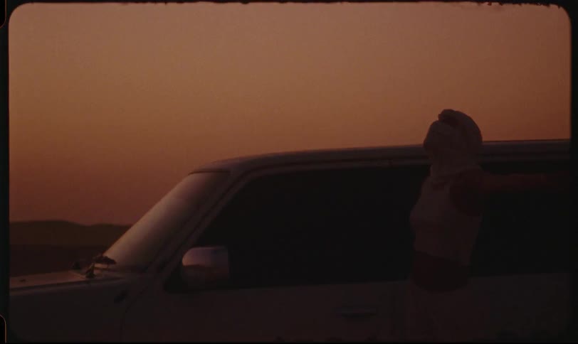 Woman leans against the outside of a pickup truck with her arms outstretched as the vehicle moves through a desert environment in the late afternoon