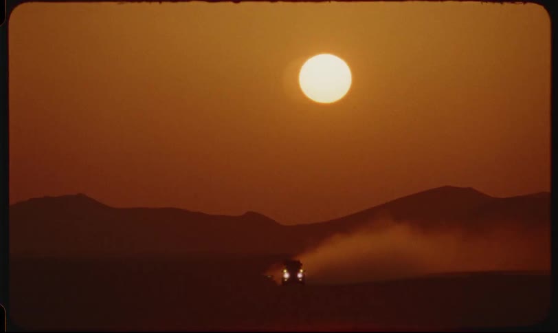 A vehicle with illuminated headlights kicks up dust as it travels across a desert landscape under a large hazy sun setting or rising in the sky