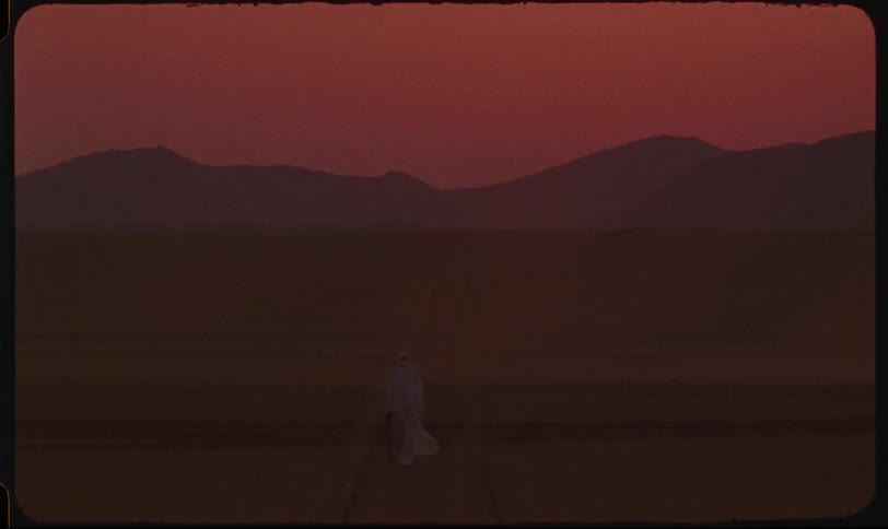 A figure draped in white walks along a track through a vast flat expanse with distant silhouetted mountains under a dusky red sky