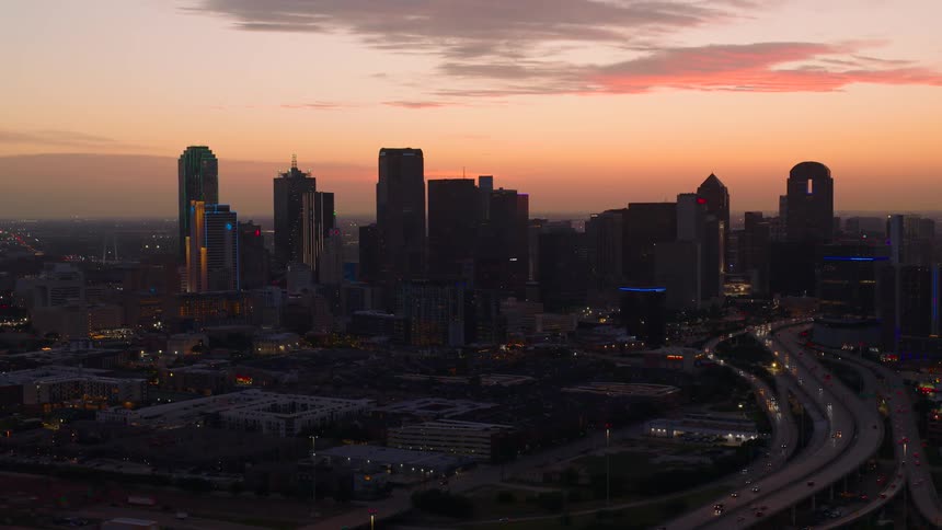 An aerial view captures the illuminated skyline of dallas texas against a vibrant sunset sky with multiple highways curving through the lower portion of the frame