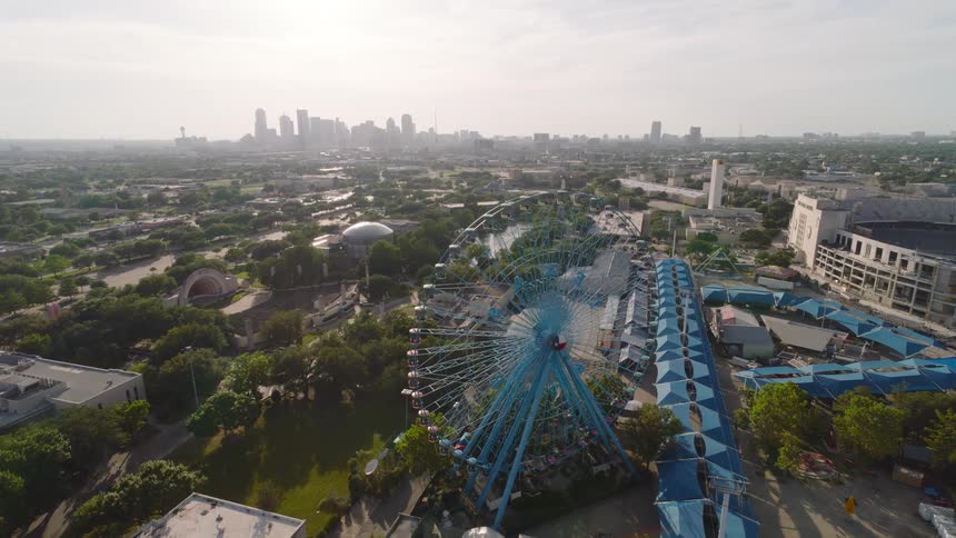 An aerial view captures a large ferris wheel at a fairground with a city skyline in the background