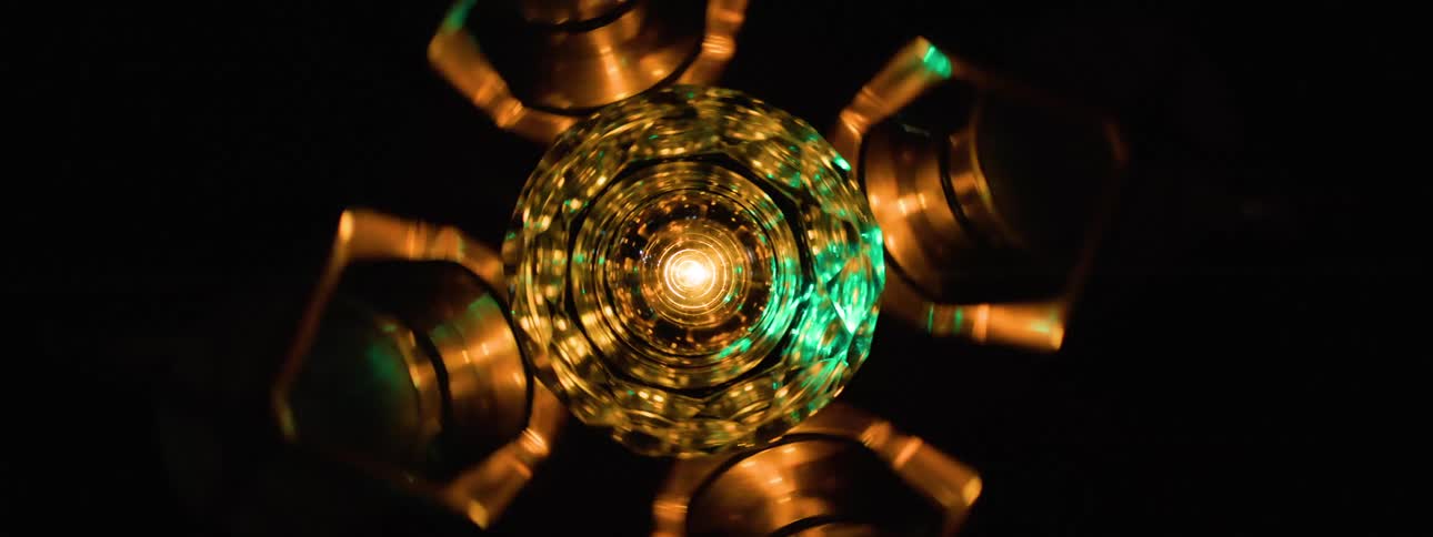 A central glowing multifaceted sphere is surrounded by metallic hexagonal arms in a dark possibly industrial or technological setting with flashing green light