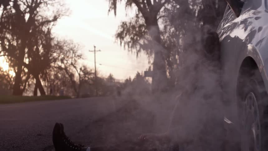 A woman is leaning against the side of  car as smoke comes it