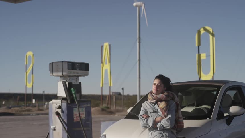 Woman sitting against the front of her car and then walking me towards the gas pump to pick up the nozzle on a clear day