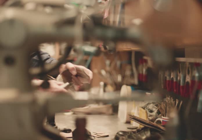 A shoemakers hands are visible working with a lightcolored material at a cluttered workbench filled with tools and shoe parts in a workshop setting