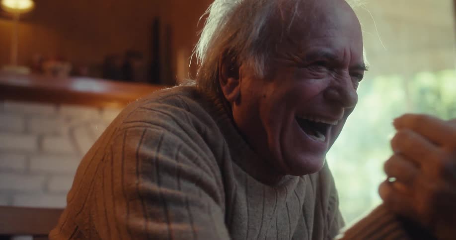 An elderly man wearing a turtleneck sweatshirt playfully shakes another man while laughing seated indoors in a chair