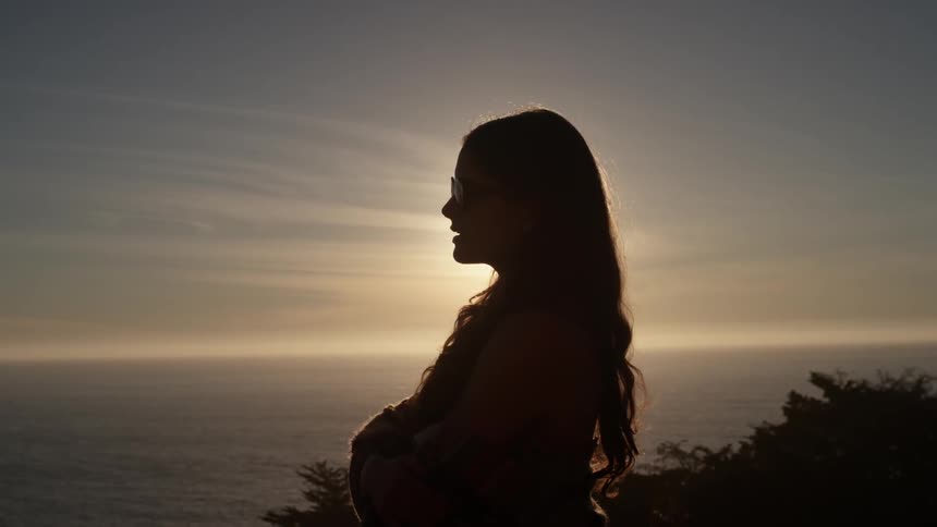 A woman stands atop a cliff looking out over the water
