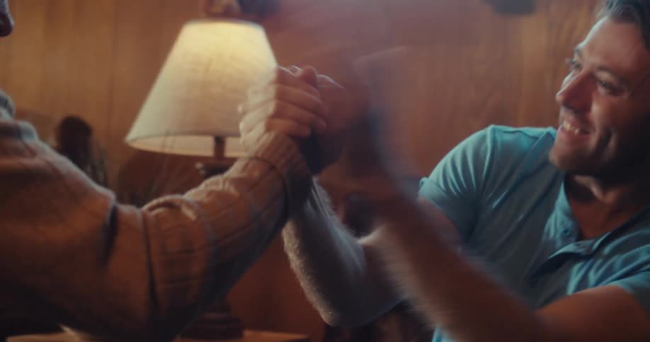 An elderly man and another man playfully perform a rehearsed handshake in a room