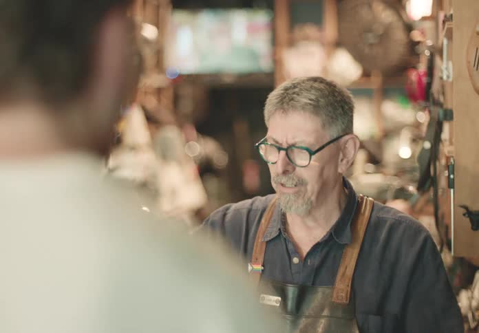 In a cluttered workshop a mature man with glasses and a leather apron is speaking to another person