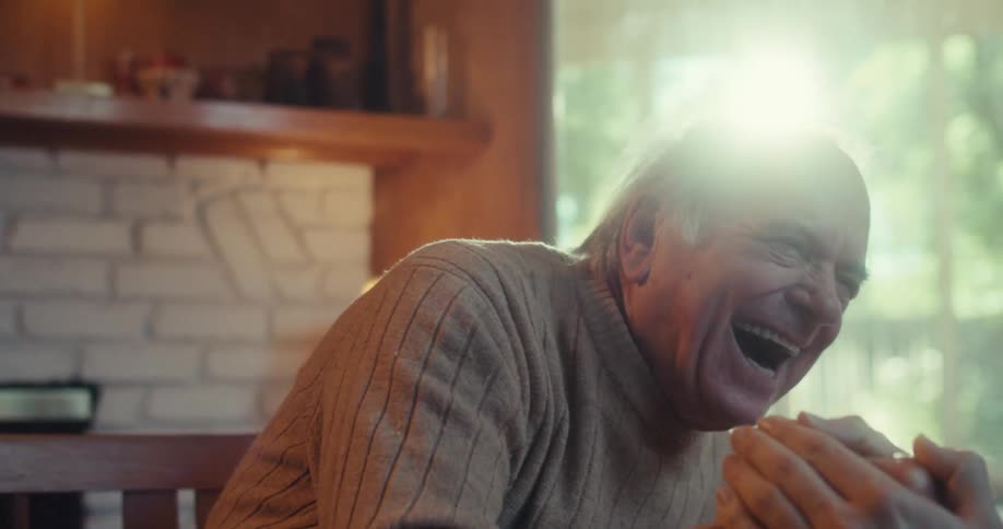 Elderly man laughs and playfully shakes hands with a younger man while sitting at home